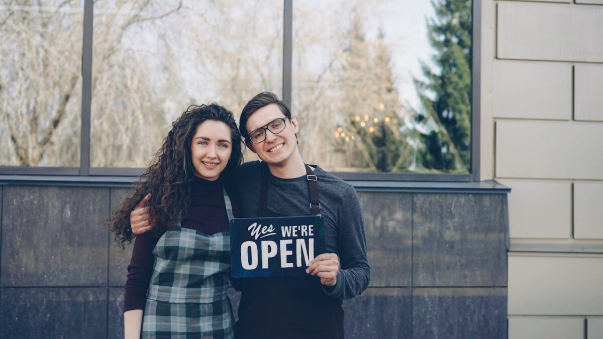 Happy small business owners embracing and displaying an open sign outdoors.